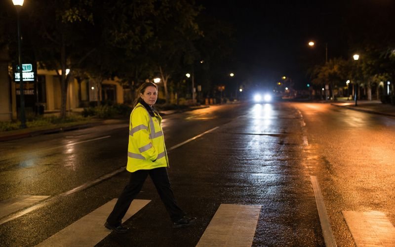 Pedestrian wearing reflective clothing crossing a California street at night with car headlights visible showing nighttime safety